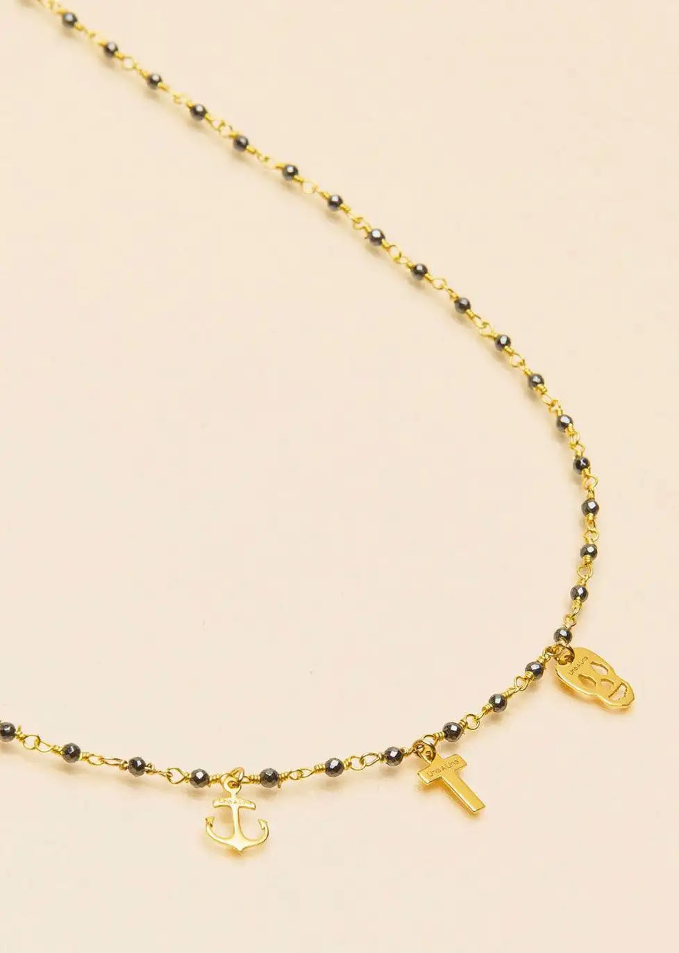 Gold chain necklace with black beads, anchor, cross, and skull charms on a neutral background.