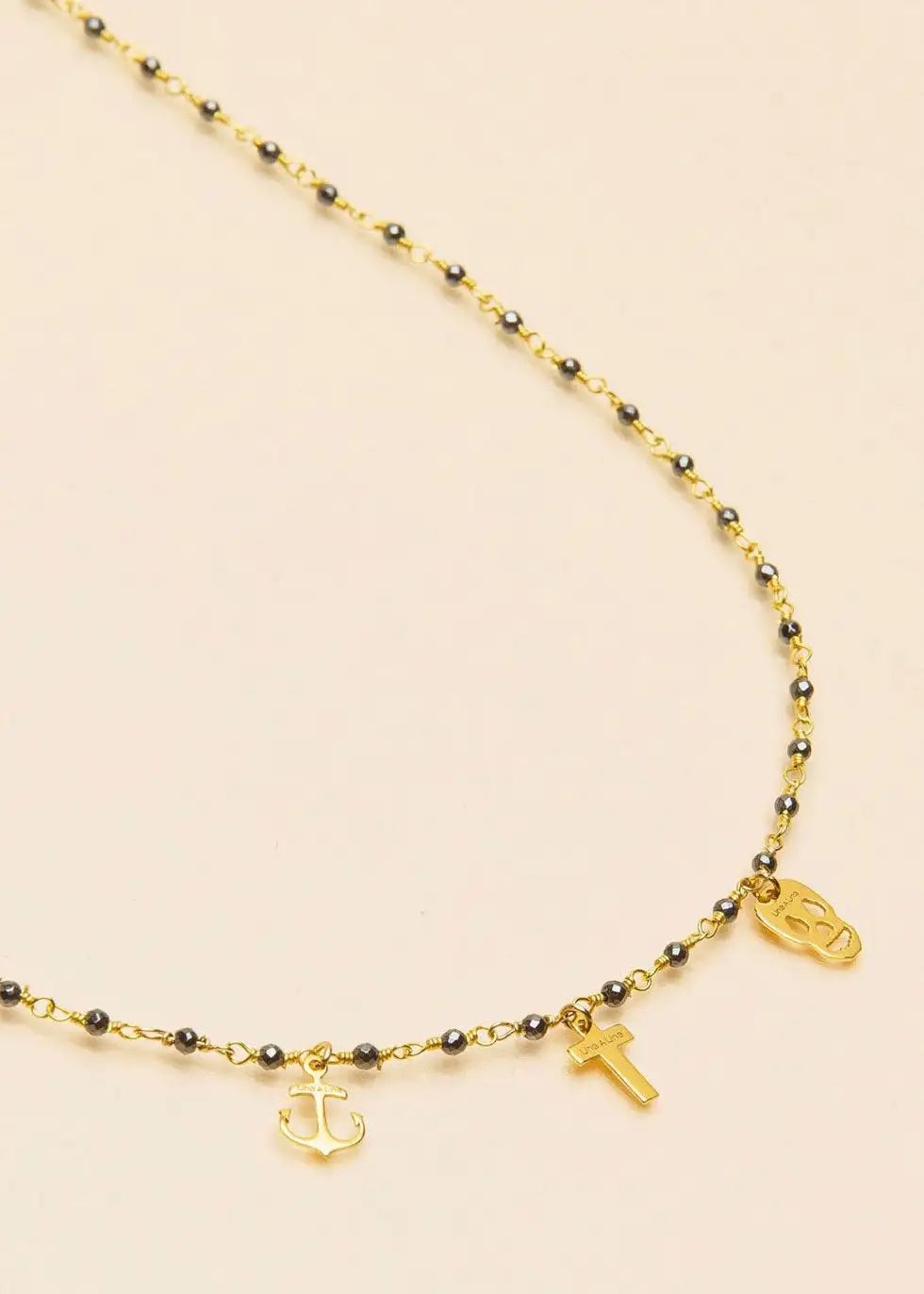 Gold chain necklace with black beads, anchor, cross, and skull charms on a neutral background.