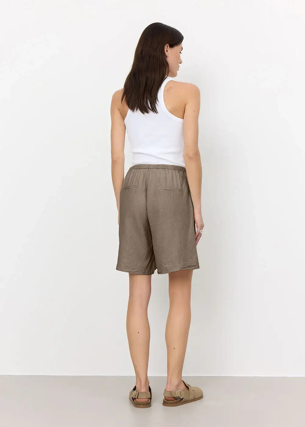 Woman in taupe linen shorts, white tank top, and sandals, seen from behind, studio setting