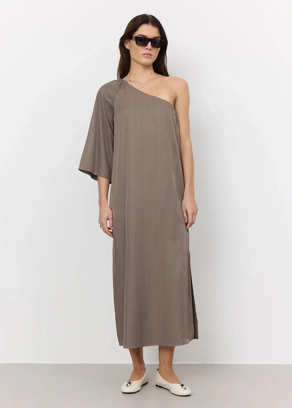 Woman wearing taupe one-shoulder maxi dress with single wide sleeve, white flats, and sunglasses