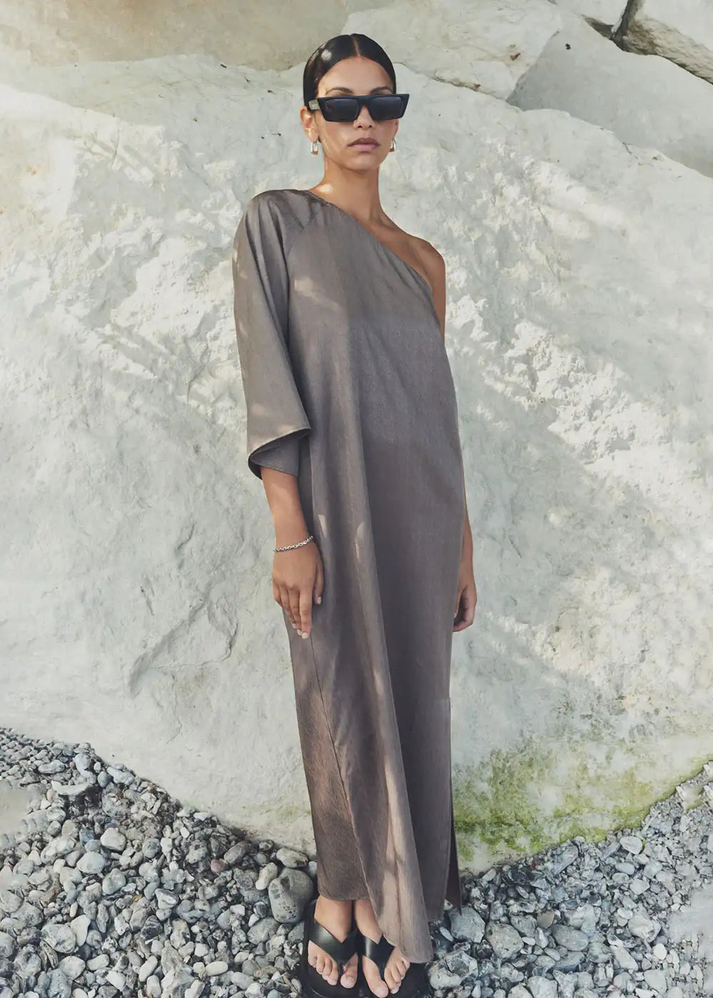 Woman in taupe one-shoulder maxi dress, sunglasses, and sandals standing on rocks outdoors