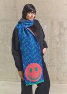 Woman wearing a colorful scarf with smiley face design against a concrete wall.