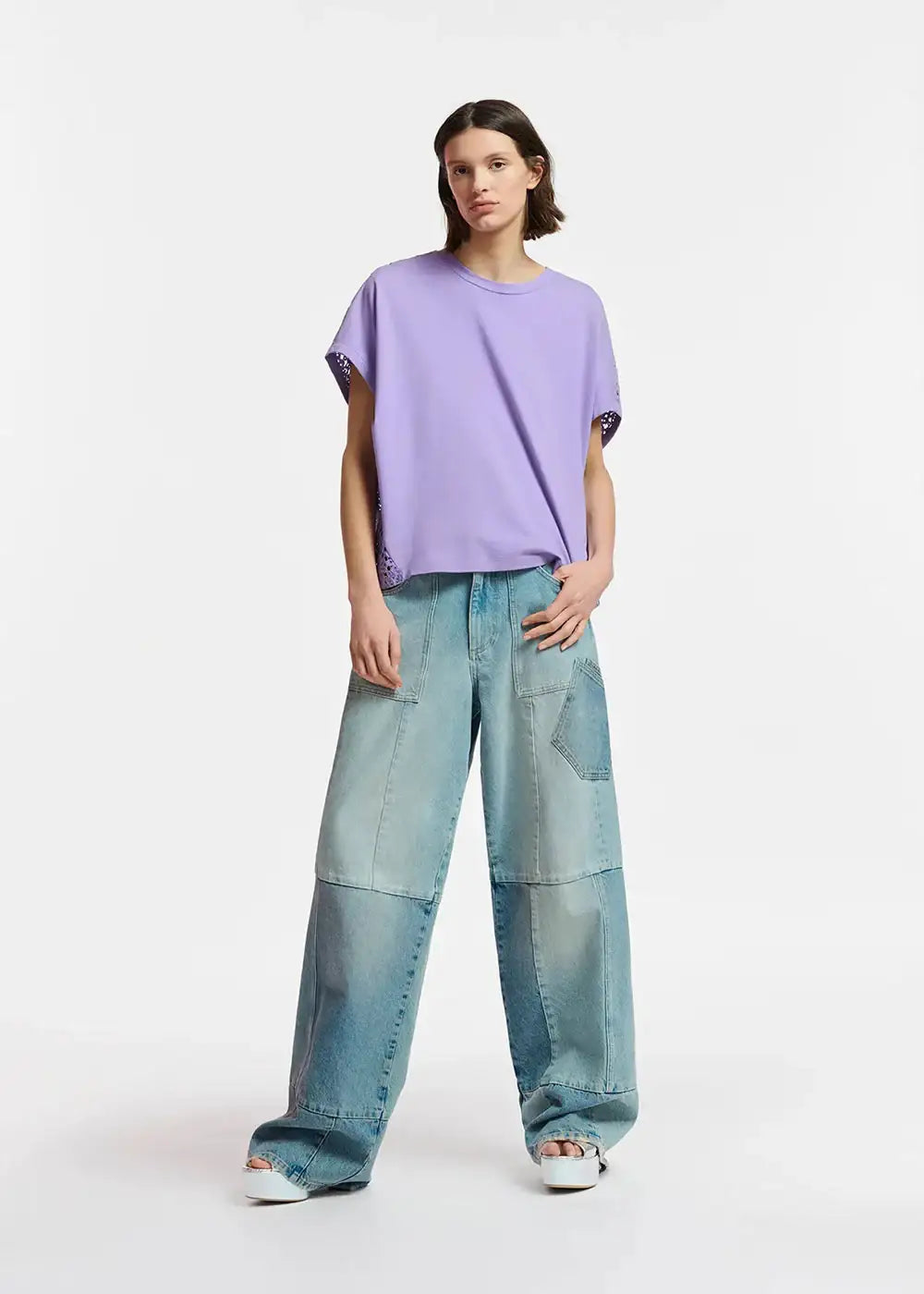Woman wearing a loose lavender t-shirt and patchwork wide-leg denim jeans on white background