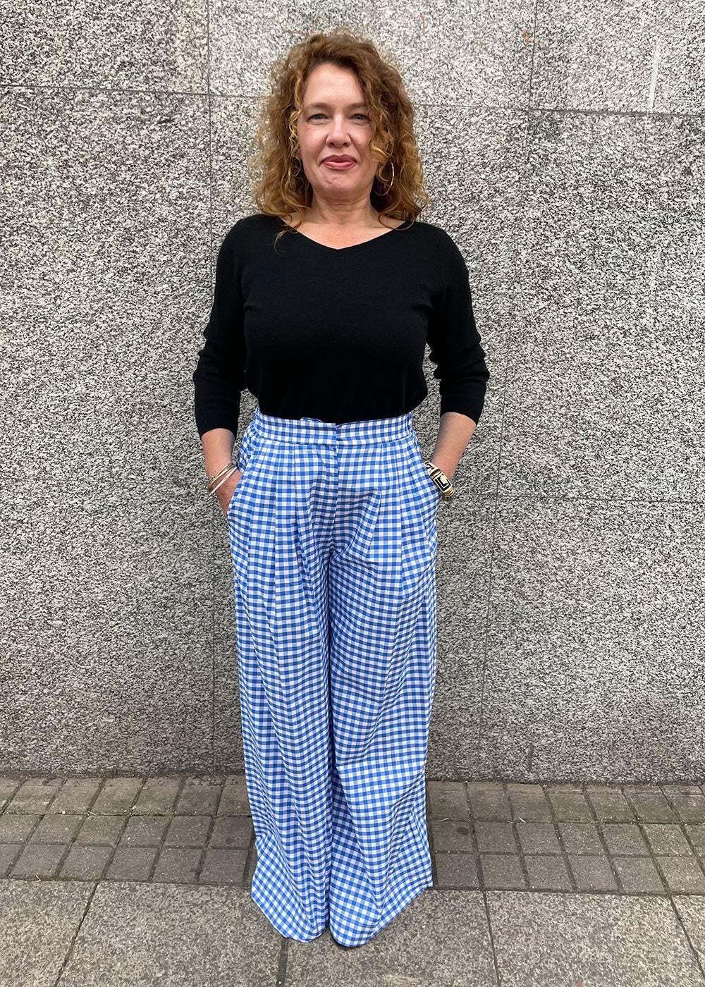 Woman in black top and blue gingham wide-leg trousers standing against a stone wall