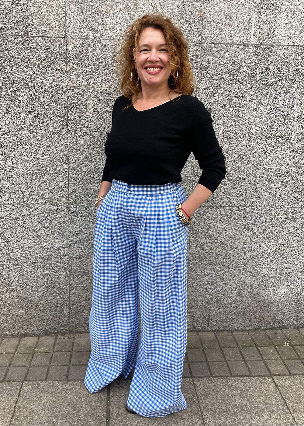 Woman in a black top and blue gingham wide-leg trousers standing against a stone wall